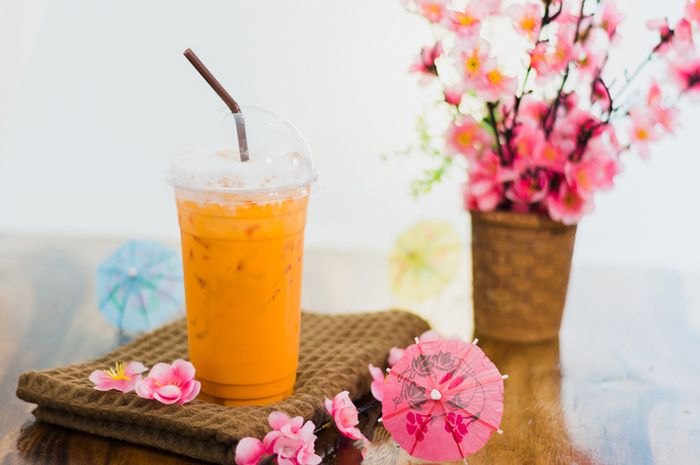 Gelas Thai Tea yang disajikan dengan es batu dan lapisan susu kental manis di atasnya, menampilkan warna oranye yang khas dan menarik, sempurna untuk hari yang panas